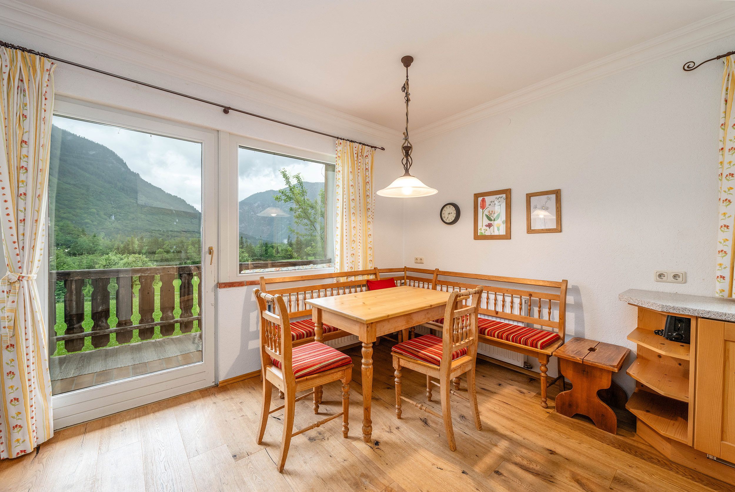 Ein Essbereich mit Holzboden, einer Bank und einem Tisch, einer Hängelampe und Blick auf Grün durch große Fenster.