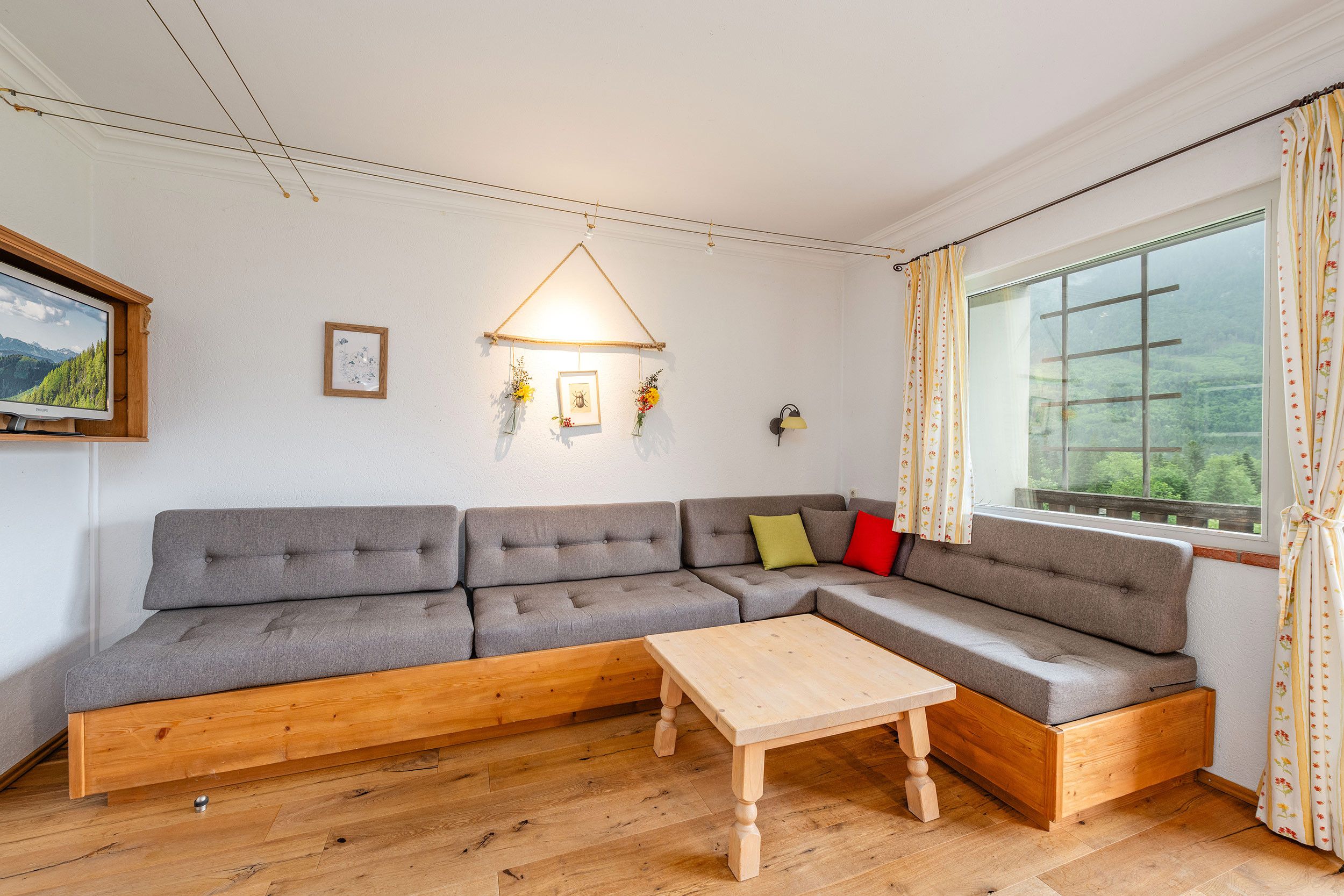 Ein modernes Wohnzimmer mit einem grauen Ecksofa, einem Holztisch und einem großen Fenster mit Vorhängen.