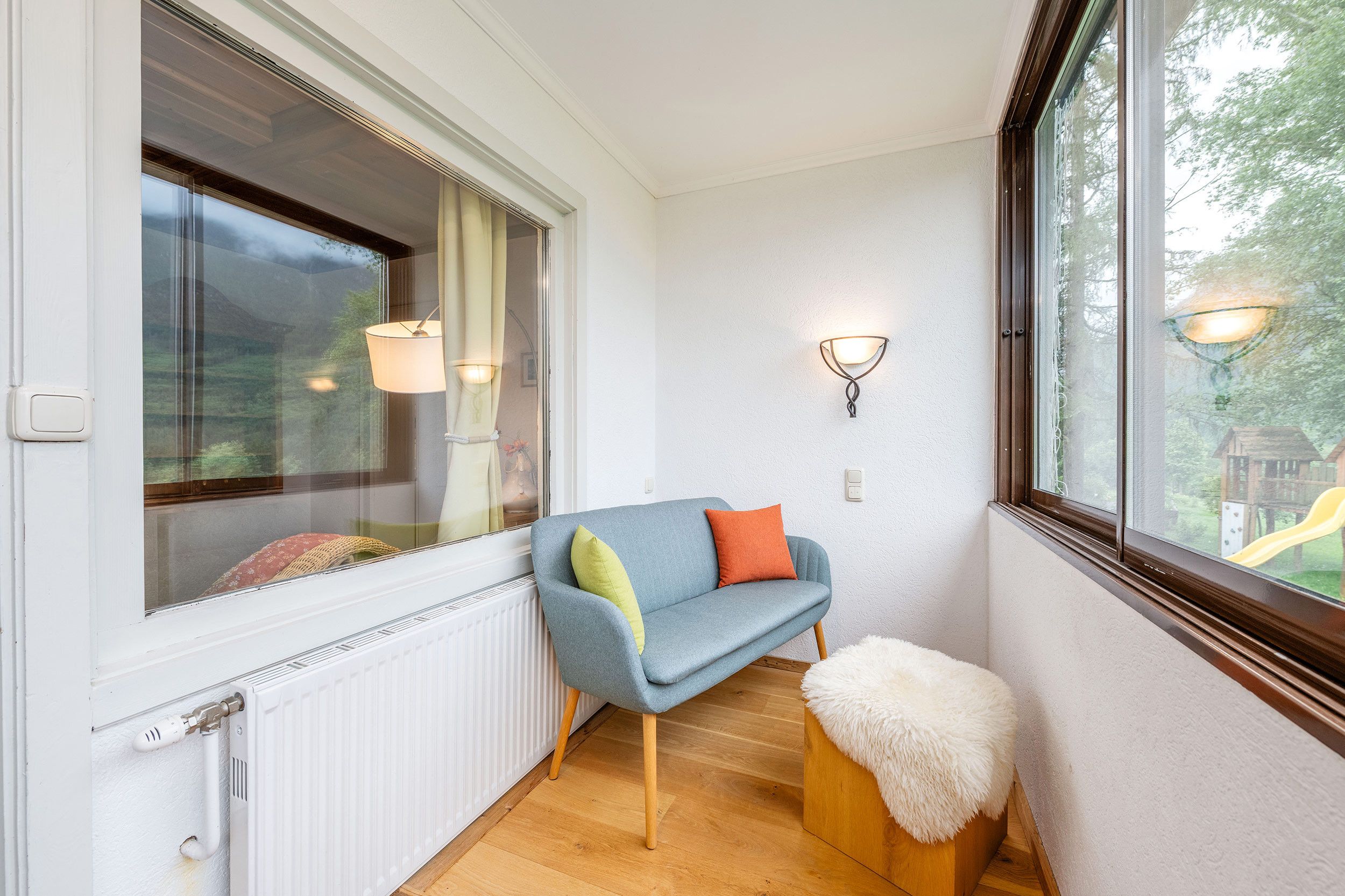 Ein Zimmer mit einem blauen Sofa, einem weißen, flauschigen Hocker und einem Fenster mit Blick auf Grünflächen.