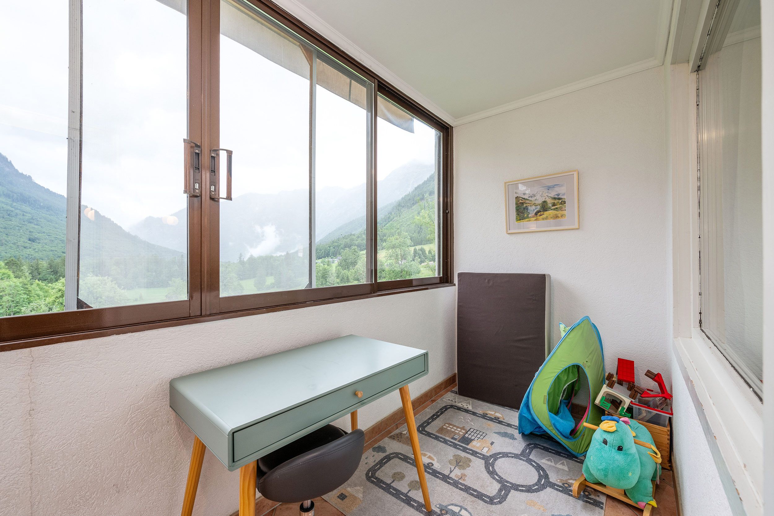 Ein Zimmer mit Schreibtisch, einem grünen Stuhl, einem Kinderspielzelt und Bergblick durch ein großes Fenster.
