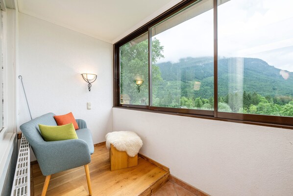 Ein Eckzimmer mit großem Fenster, einem blauen Sessel, einem kleinen Hocker und einer Wandleuchte.