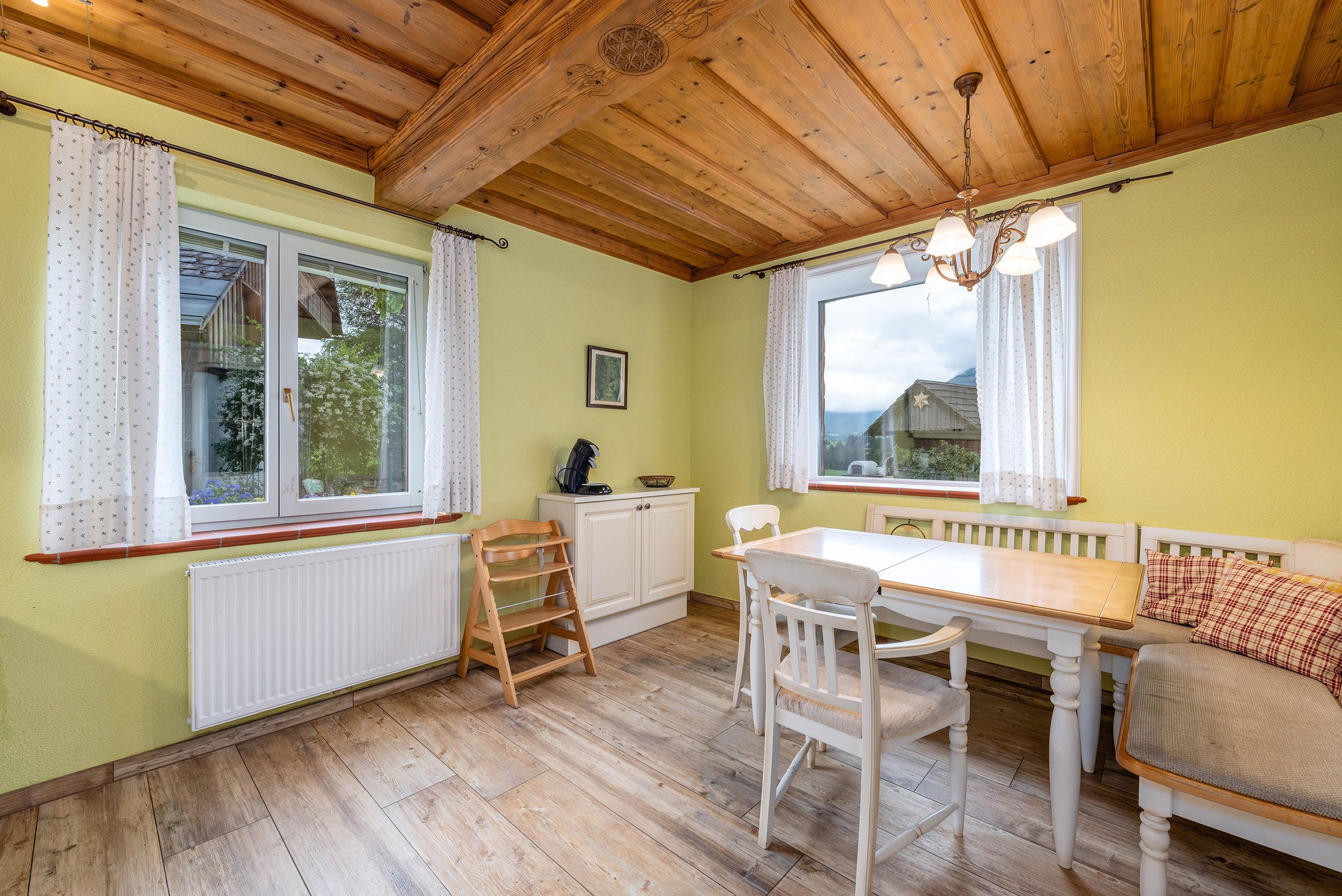 Ein Zimmer mit Holzboden, einem Esstisch mit Stühlen, einer Bank, einem Heizkörper und einem Fenster mit Vorhängen.