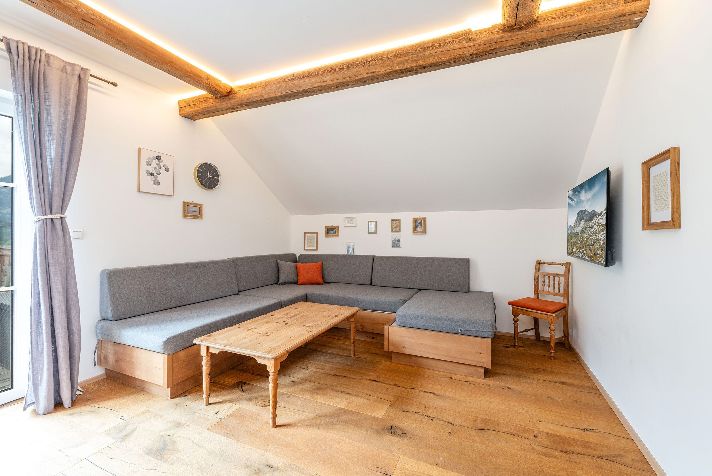 Ett modernt sovrum i loftstil med ett sluttande tak, en grå hörnsoffa, en träbänk och en trästol.