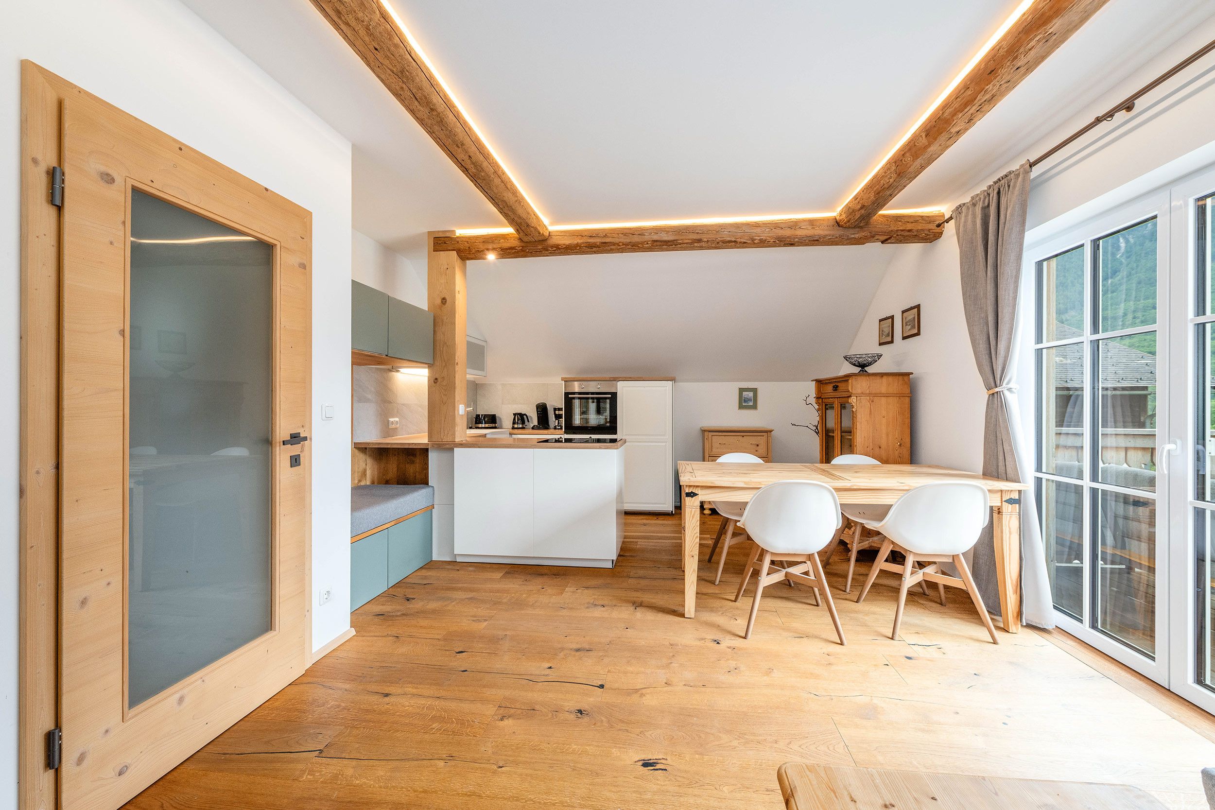 Una cucina moderna con travi in legno, un'area pranzo con tavolo e sedie, e un'ampia finestra con tende.
