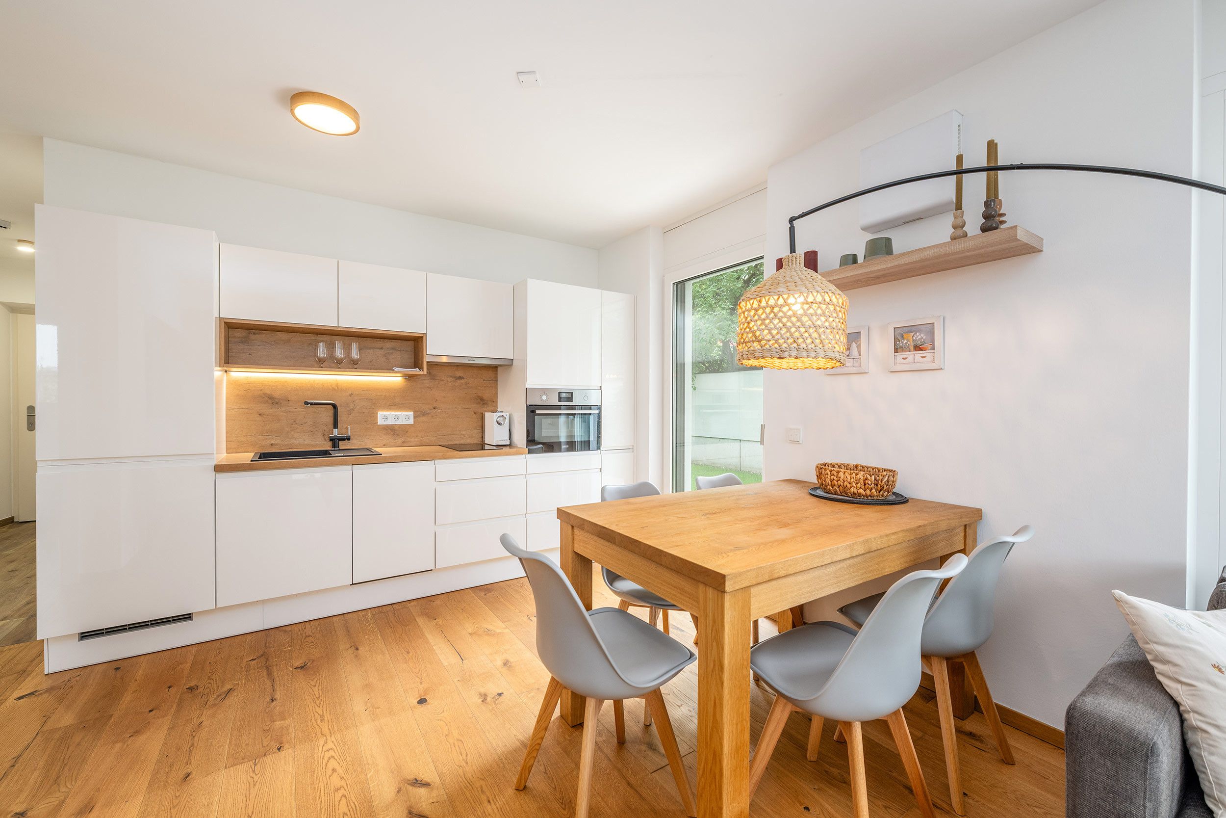 Una cocina moderna con armarios blancos, encimera de madera y zona de comedor con mesa de madera y sillas grises.