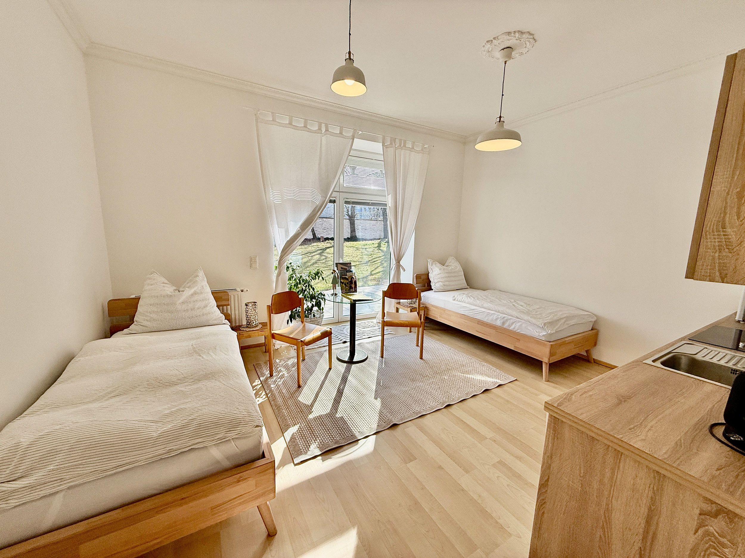 Ein kleines Zimmer mit zwei Einzelbetten, einem kleinen Tisch, Stühlen, einer Küchenzeile und einem großen Fenster.