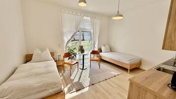 Ein kleines Zimmer mit zwei Einzelbetten, einem kleinen Tisch, Stühlen, einer Küchenzeile und einem großen Fenster.