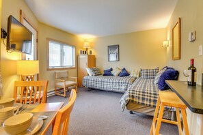 Une chambre à coucher de taille réduite, comprenant un lit, un bureau, une chaise, une petite table et une fenêtre avec des stores.