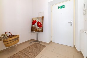 Un pasillo con una puerta blanca, un cuadro de flores rojas, una cesta de mimbre y una mesita con tapa de vidrio.