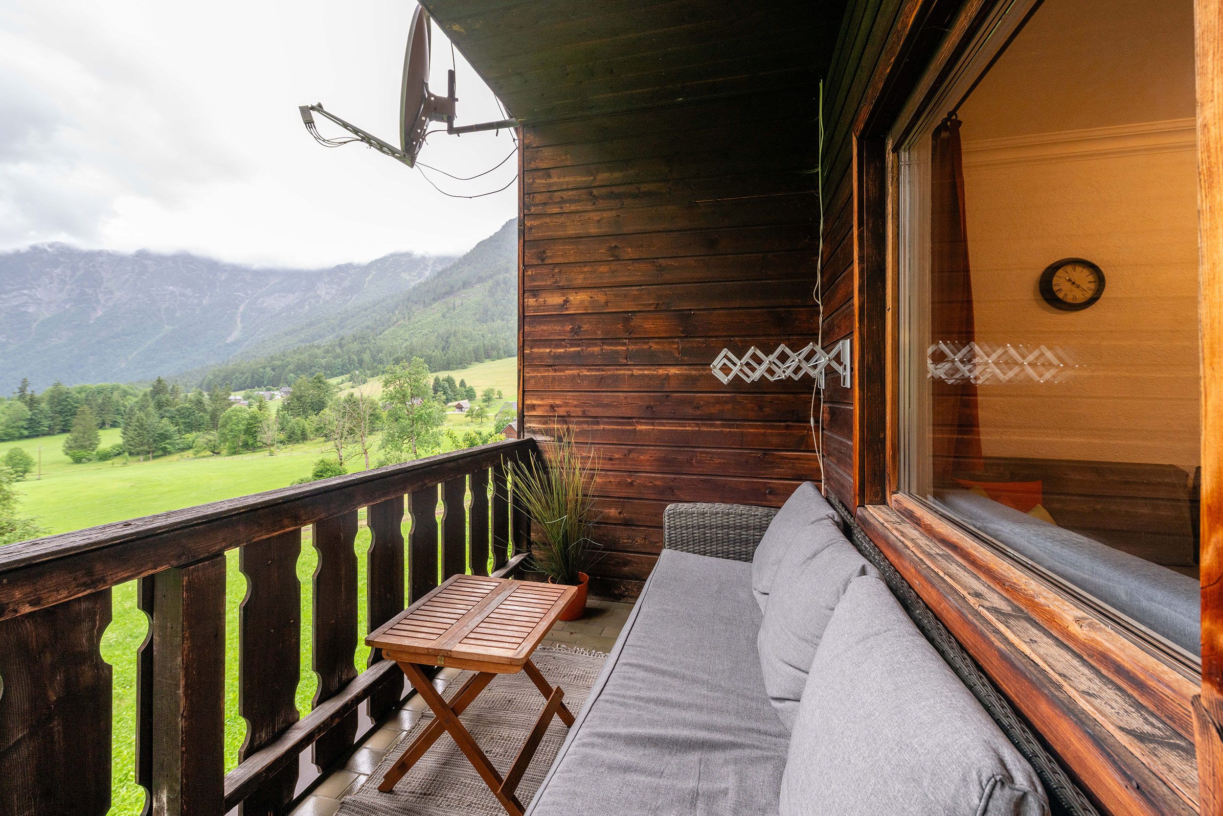 A wooden balcony with a sofa and table, overlooking a green landscape and mountains.