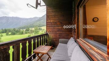 A wooden balcony with a sofa and table, overlooking a green landscape and mountains.