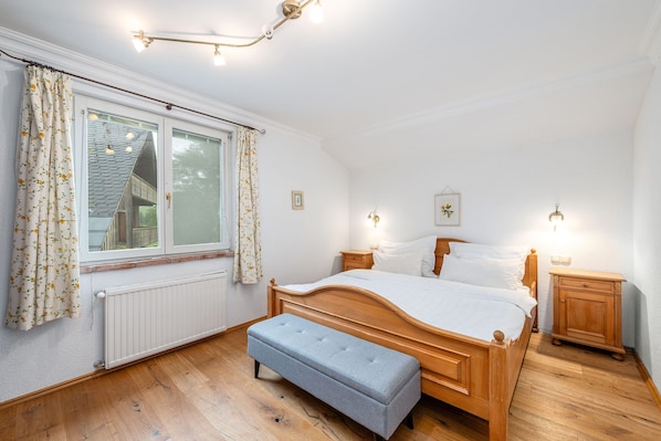 Ein Schlafzimmer mit einem hölzernen Bett, einer Bank, einem Nachttisch und einem Fenster mit Vorhängen.