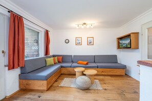 Ein gemütliches Wohnzimmer mit einer Eckcouch, einem Holztisch und einem großen Fenster mit Vorhängen.