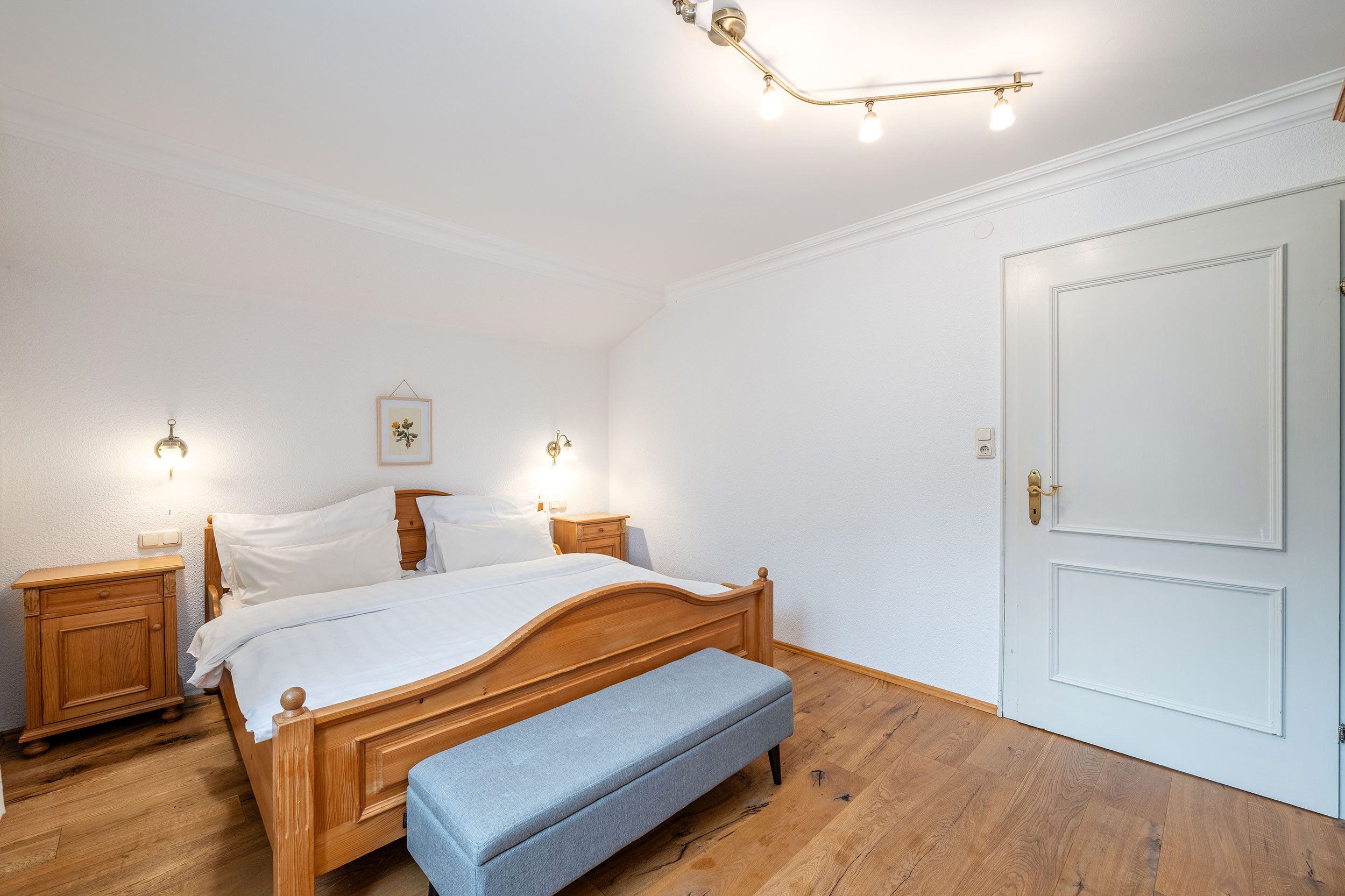 Une chambre à coucher avec un lit en bois, des tables de chevet, un banc, une porte et un luminaire au plafond.