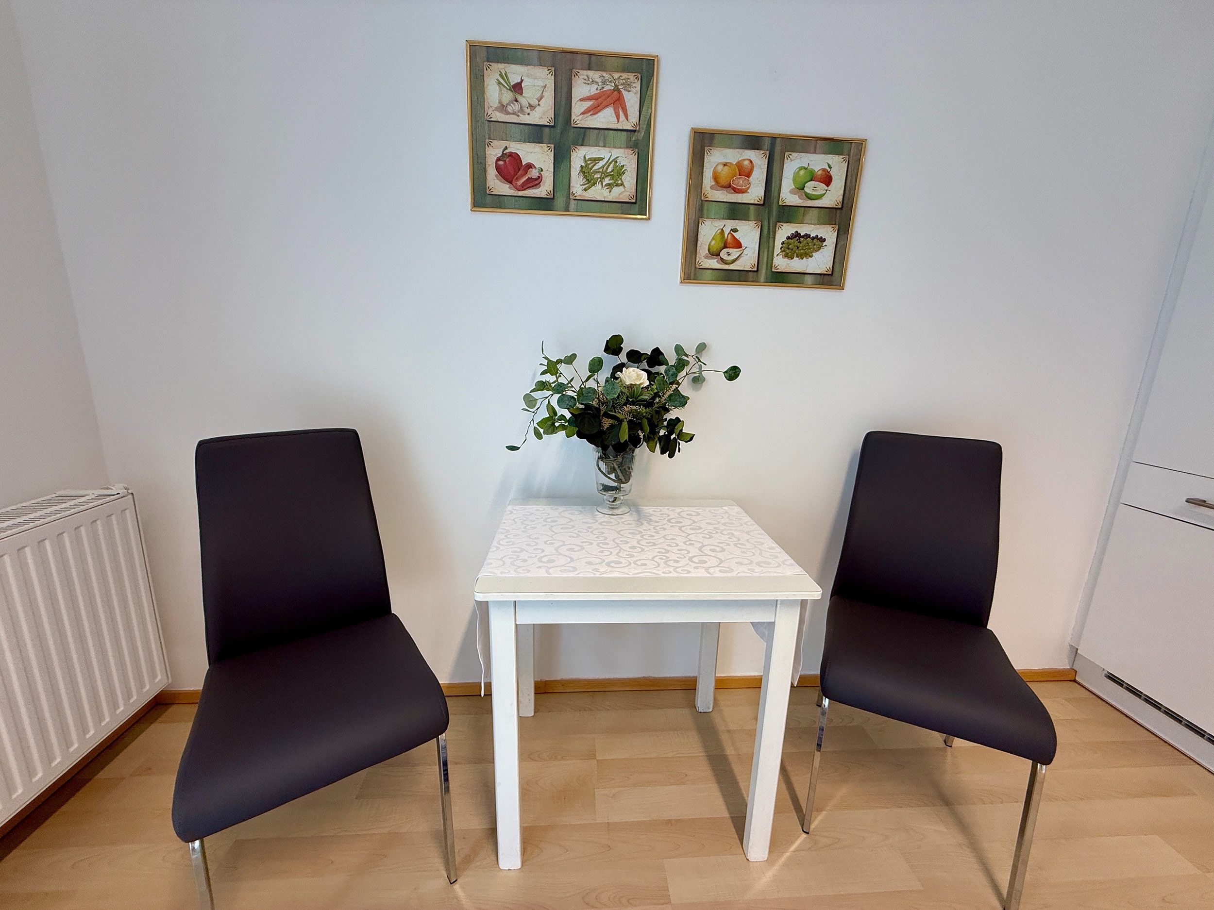 Ein Zimmer mit zwei schwarzen Stühlen, einem weißen Tisch und einer Vase mit Blumen. An der Wand befinden sich gerahmte Bilder.