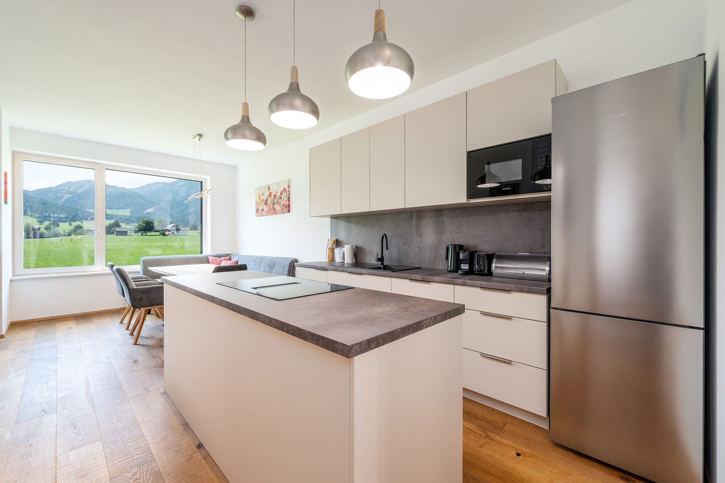A modern kitchen with a central island, stainless steel appliances, and a view of a green landscape through large windows.
