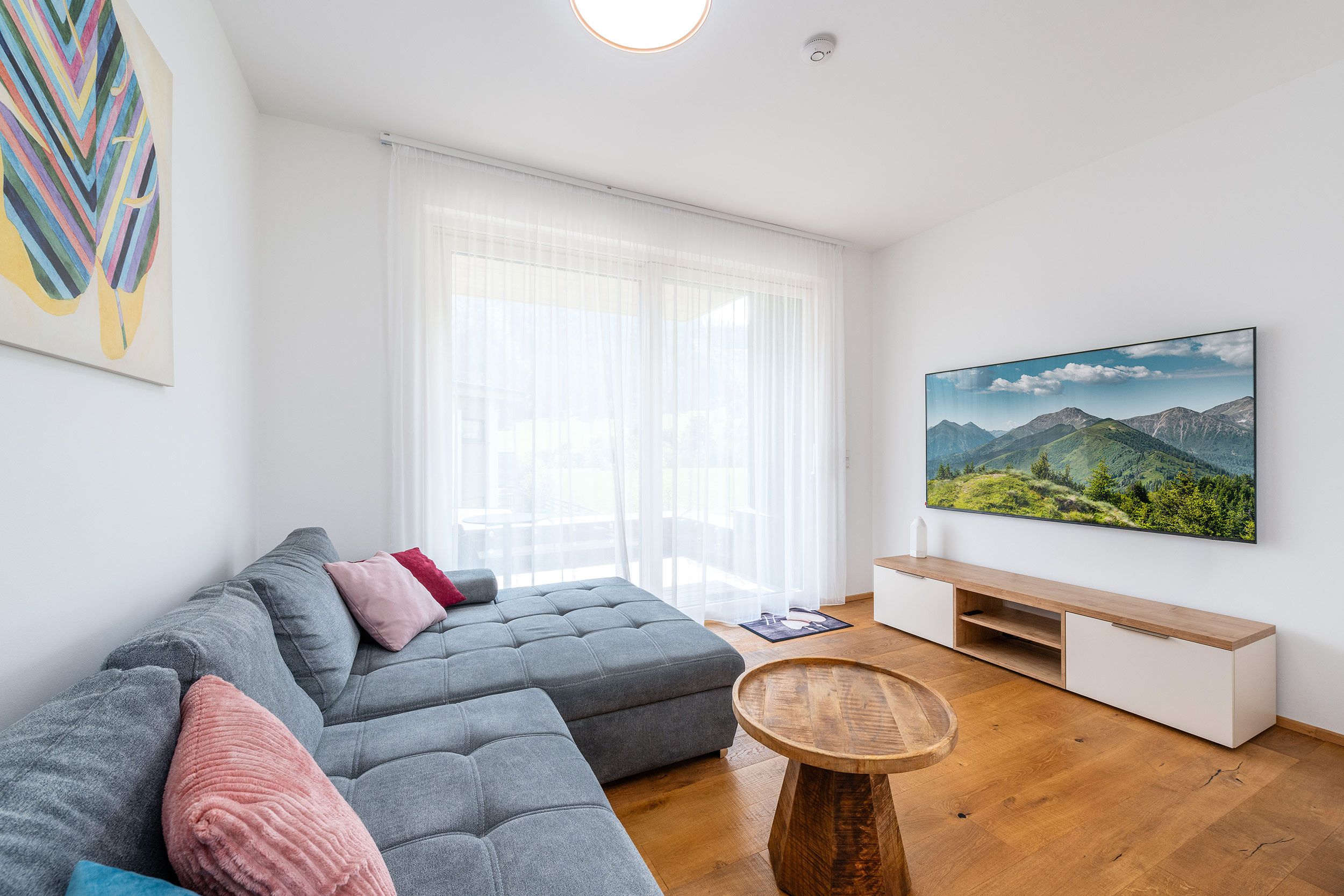 A modern living room with a grey sectional sofa, a round wooden coffee table, and a large flat-screen TV mounted on the wall.