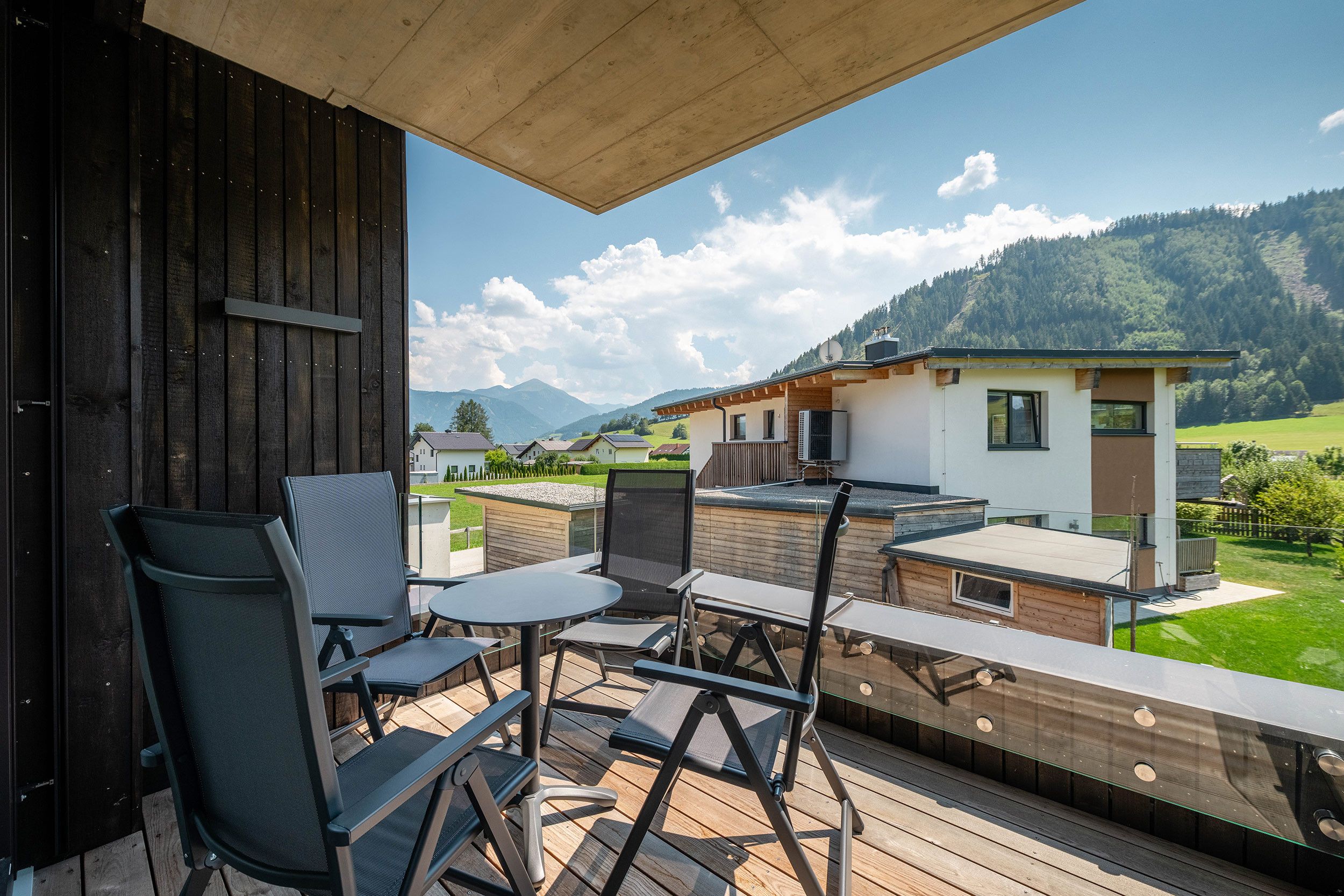 A wooden deck with outdoor furniture, a view of a mountain landscape, and a modern building in the background.