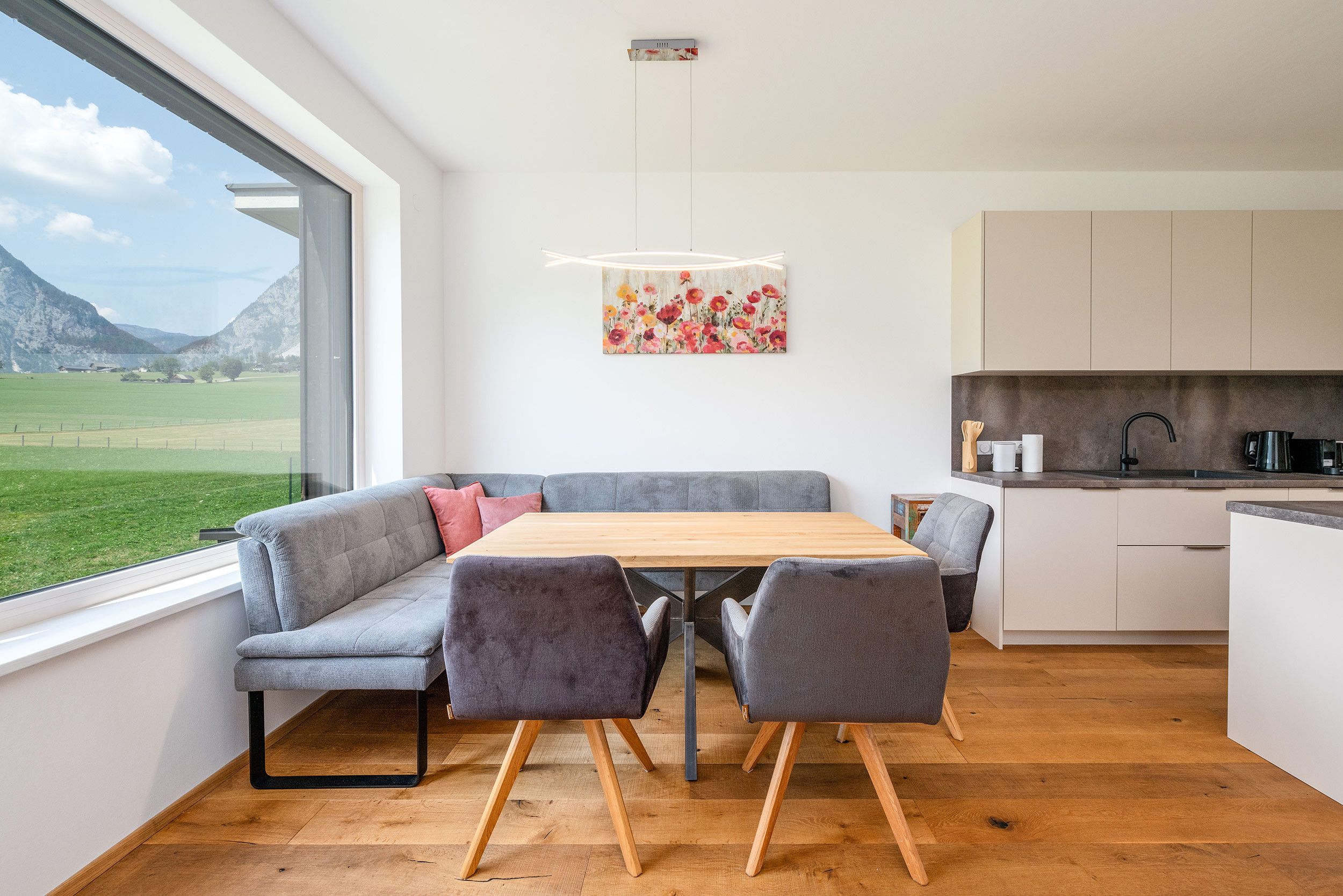 Een moderne keuken met een houten tafel en grijs gestoffeerde zitplaatsen, uitzicht op een groen landschap door een groot raam, en een schilderij aan de muur.