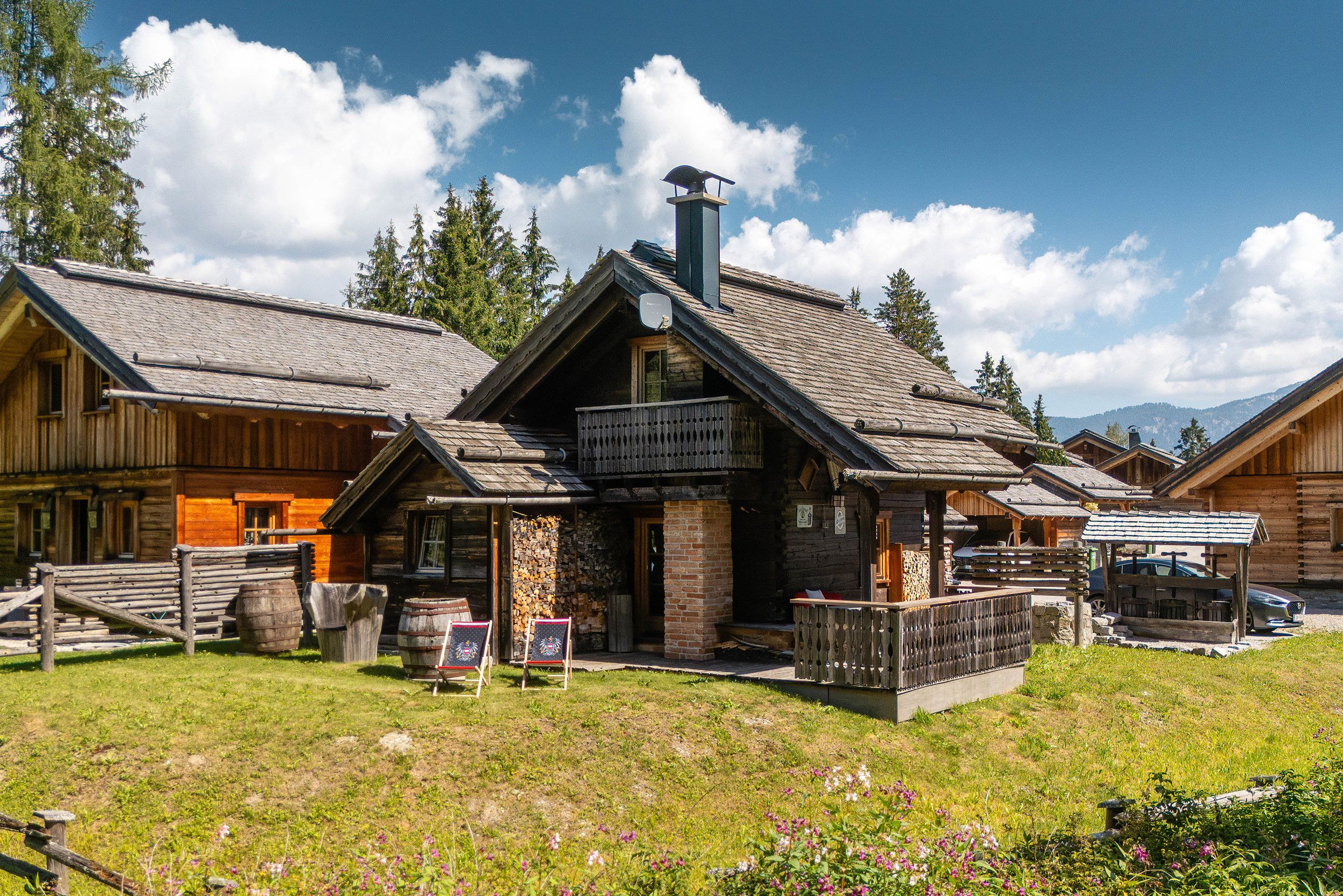 Un chalet rustique doté d’une cheminée imposante, d’une façade en bois et d’une pelouse verdoyante à l’avant, délimitée par une clôture en bois.
