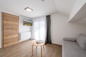 A modern living room with a wooden door, a radiator, a table with magazines, a grey sofa, and a large window with sheer curtains. - Alpenpanorama Assach 2 (Aich-Assach)