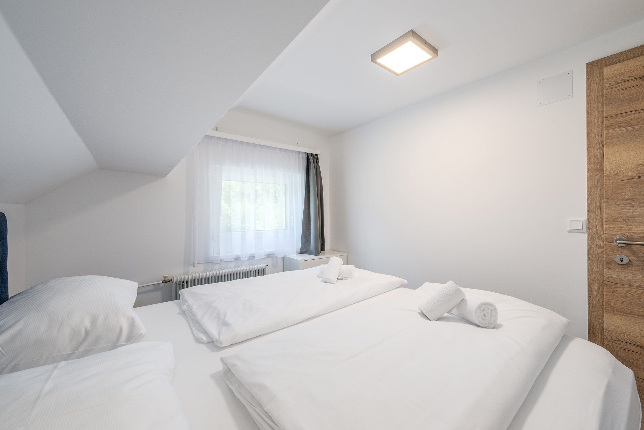 A hotel room with two beds, a window with sheer curtains, a radiator, and a wooden door.