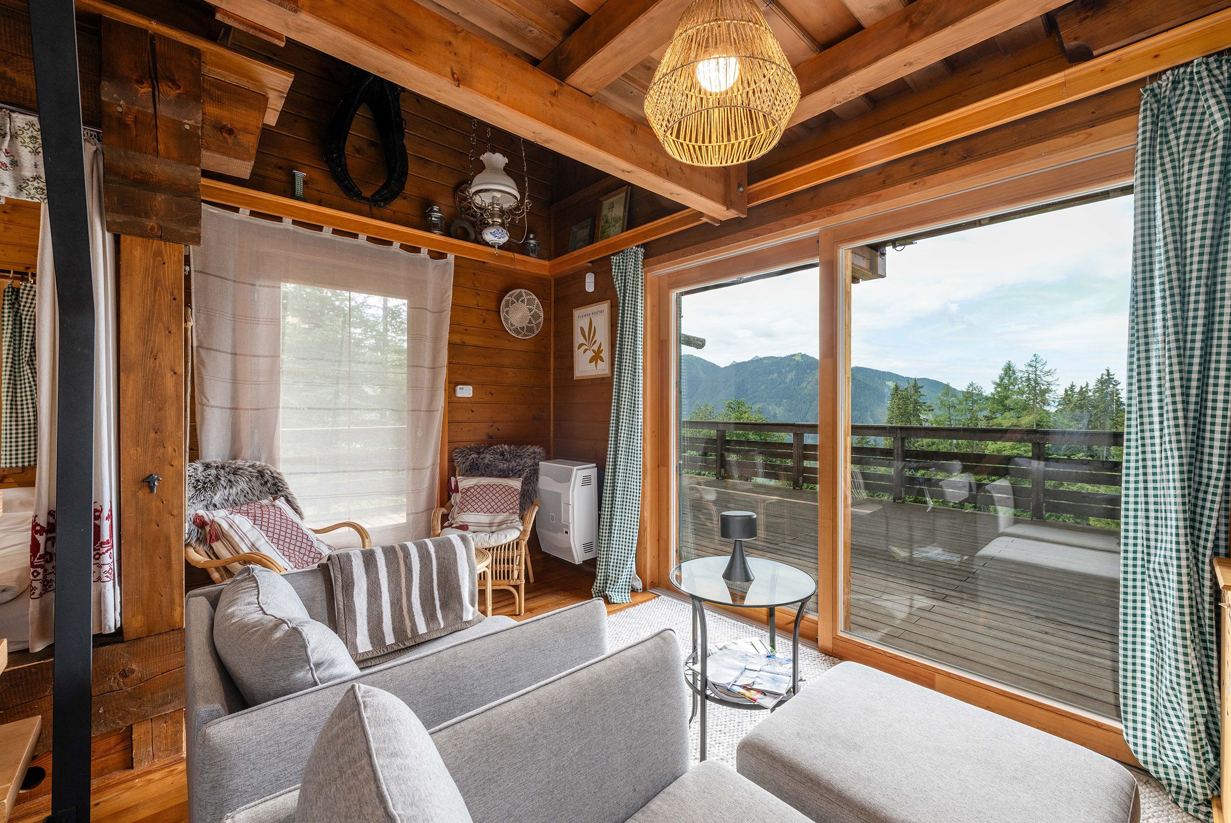 A cozy cabin interior with a sofa, a small table, and a large window offering a view of a wooden deck and mountains.