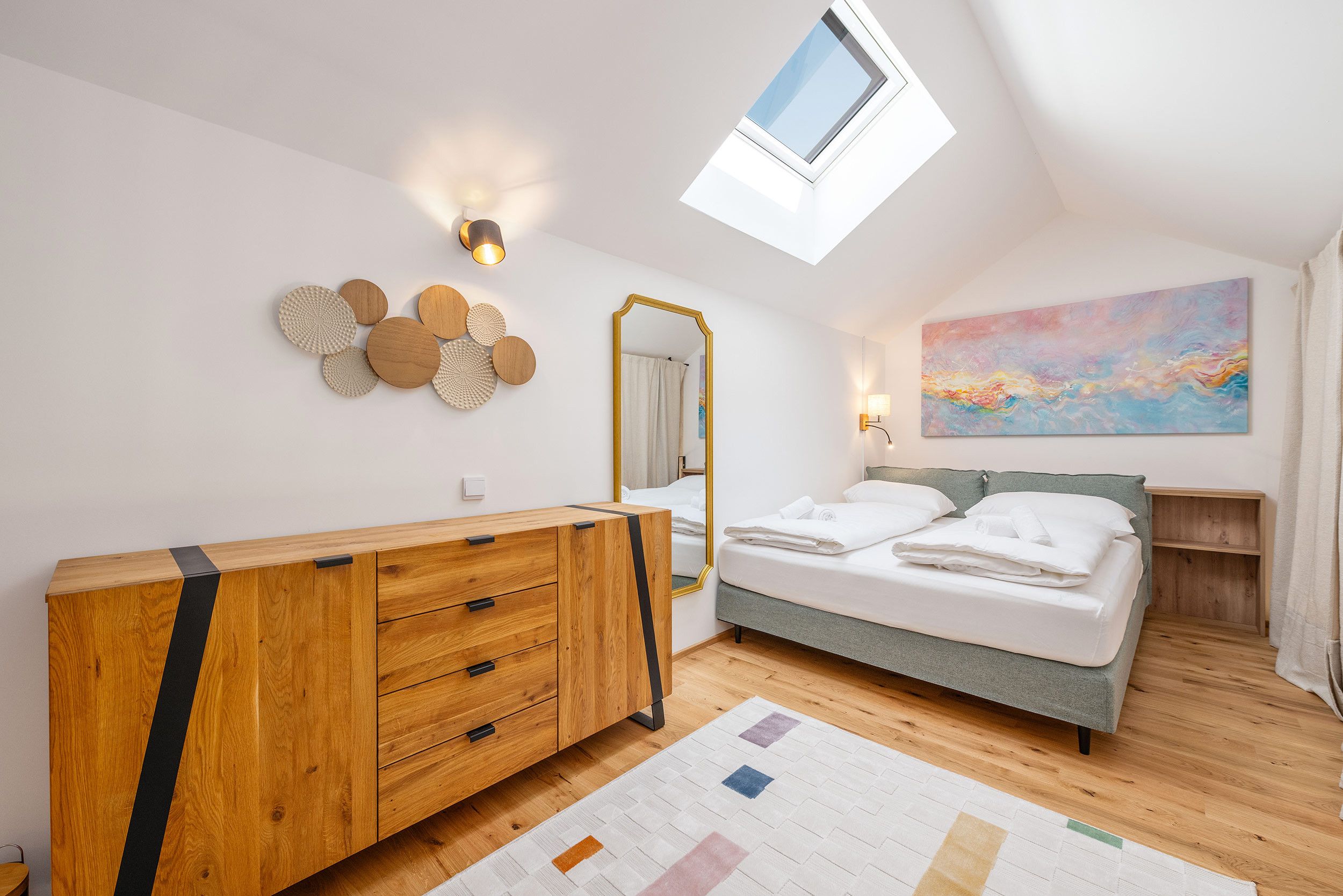 Une chambre à coucher avec une commode en bois, un lit recouvert de draps blancs, un puits de lumière, un miroir fixé au mur et un tableau abstrait aux couleurs vives.