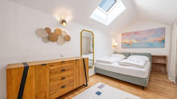 Une chambre à coucher avec une commode en bois, un lit recouvert de draps blancs, un puits de lumière, un miroir fixé au mur et un tableau abstrait aux couleurs vives.