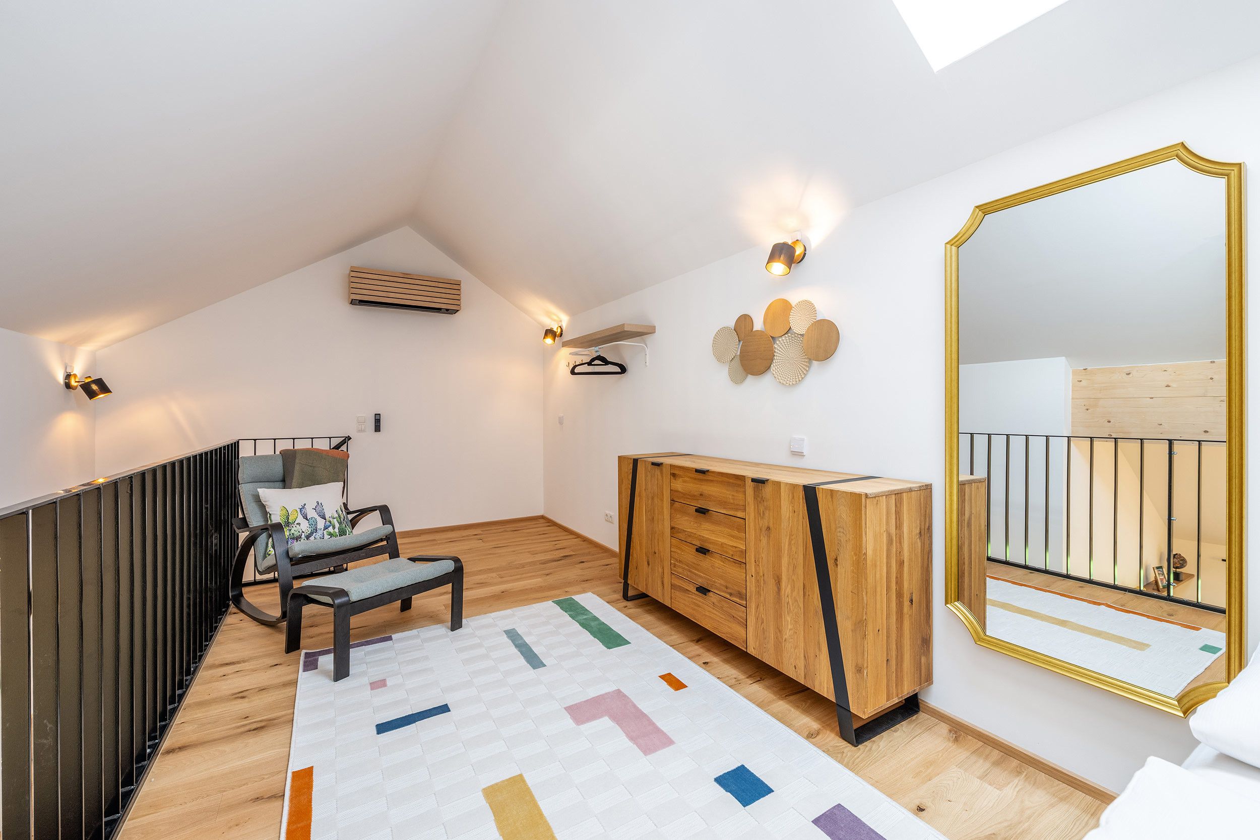 Ein modernes Schlafzimmer im Loft-Stil mit schrägverdeckter Decke, einer Holzkommode, einem Sessel mit Kissen, einem farbenfrohen Teppich und einem großen Spiegel mit goldener Rahmung.