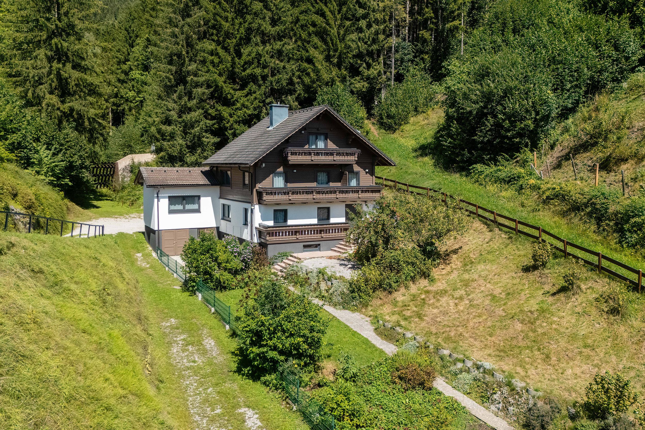 Eine zweigeschossige Chalet mit Balkon, umgeben von üppiger Vegetation und einem bewaldeten Hügelhang.