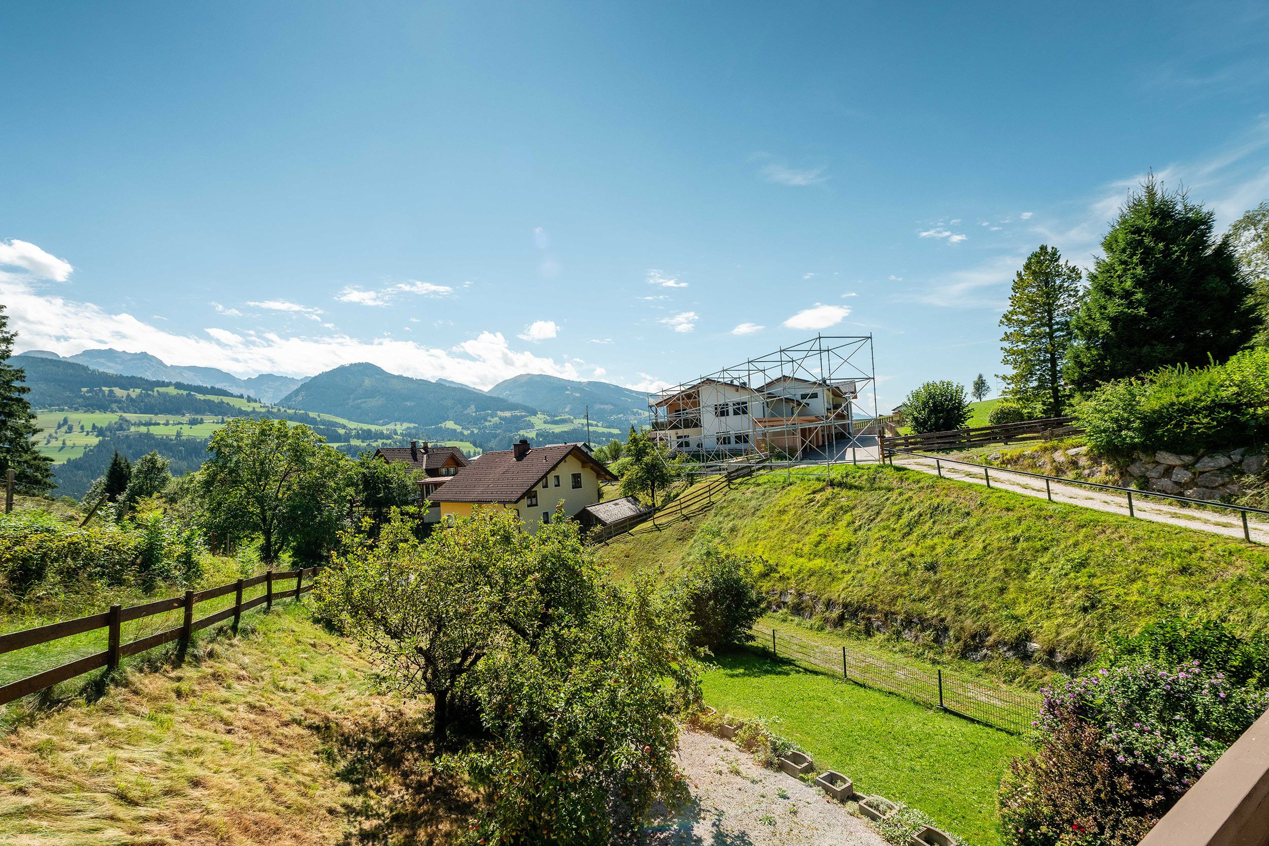 A rural landscape with a house on a hillside, surrounded by greenery and mountains in the background.