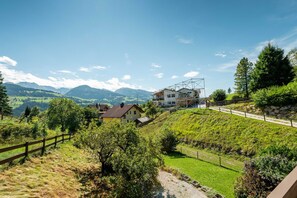 Ländliche Landschaft mit einem Haus an einem Hügel, umgeben von viel Grün und Bergen im Hintergrund.