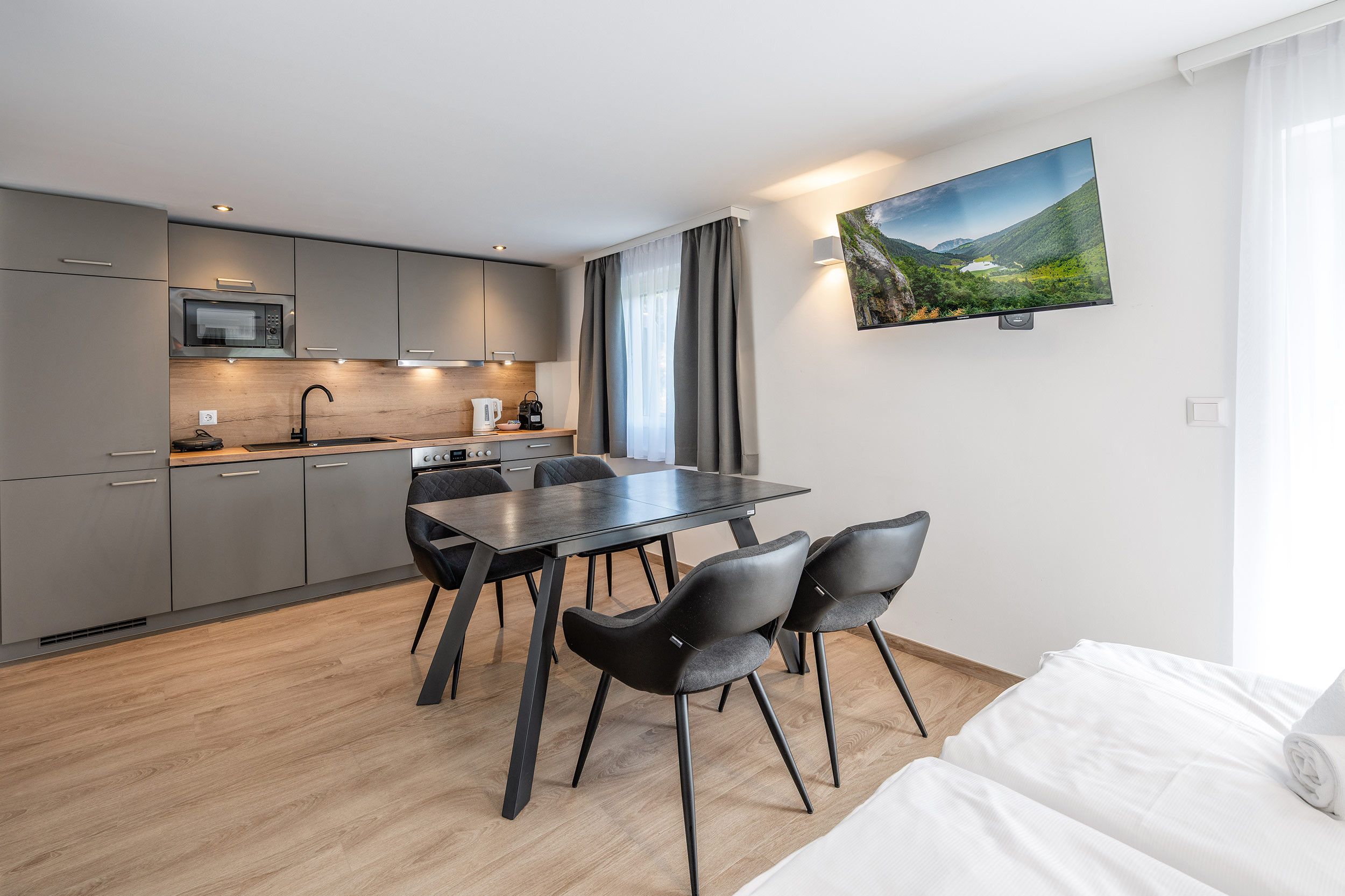 A modern kitchen with a dining area, a flat-screen TV mounted on the wall, and a bed with white linens.