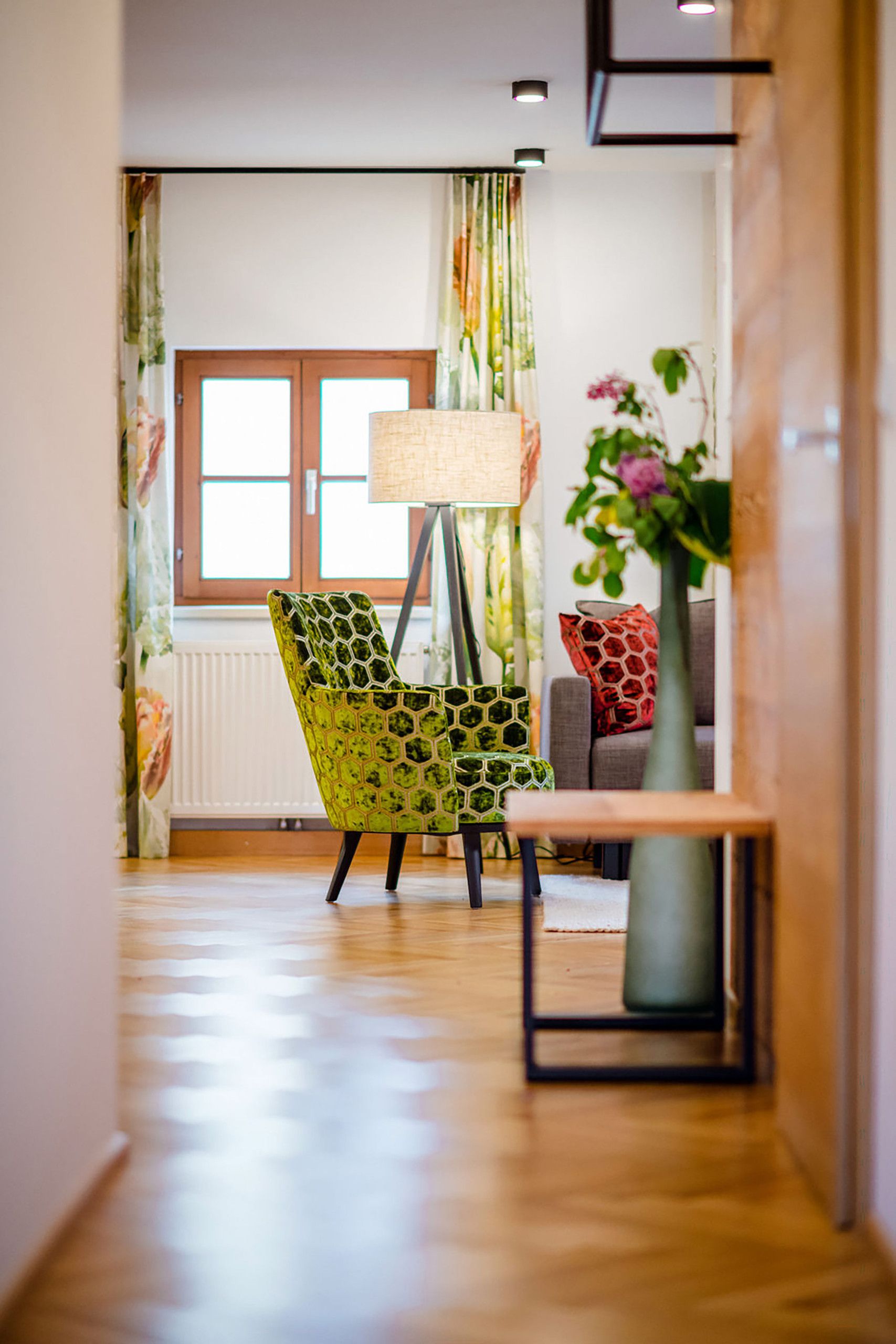 Un corridoio con una poltrona verde decorata con un motivo esagonale, una lampada da terra, una finestra con tende e un vaso con fiori.