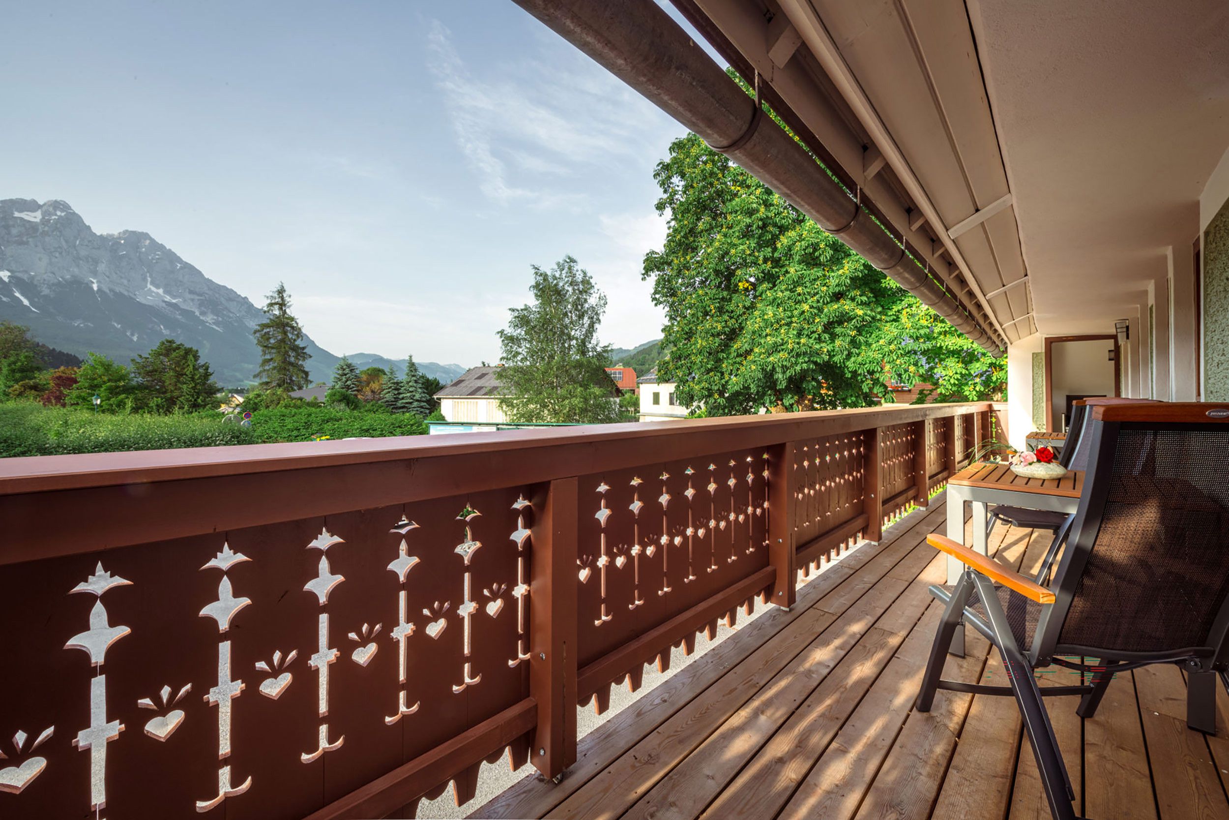 Un terrazzo in legno con ringhiera, un tavolo con sedie e vista sulle montagne e alberi.