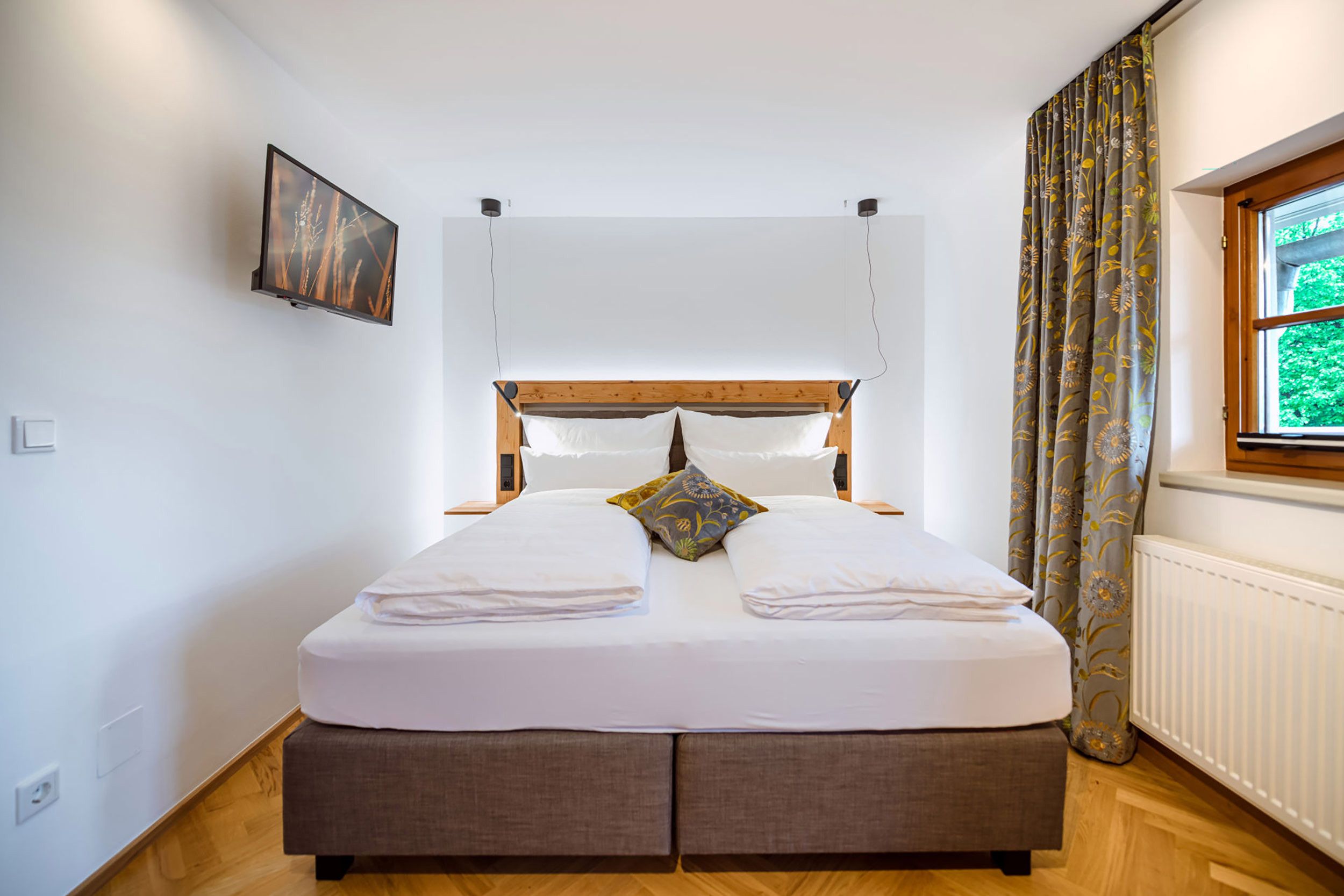 Un lit bien fait, avec une tête de lit, un téléviseur fixé au mur et une fenêtre avec des rideaux.