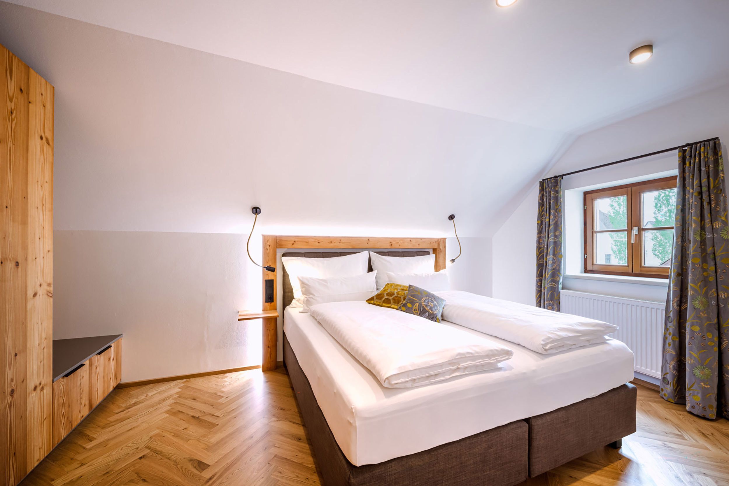 A bedroom with a wooden headboard, a bed with white bedding, a window with curtains, and a radiator.