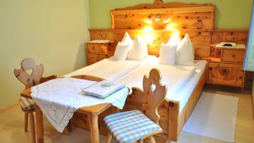A wooden bed with a headboard, two bedside tables, a nightstand, a wooden chair with a checkered cushion, and a small table with a white cloth.