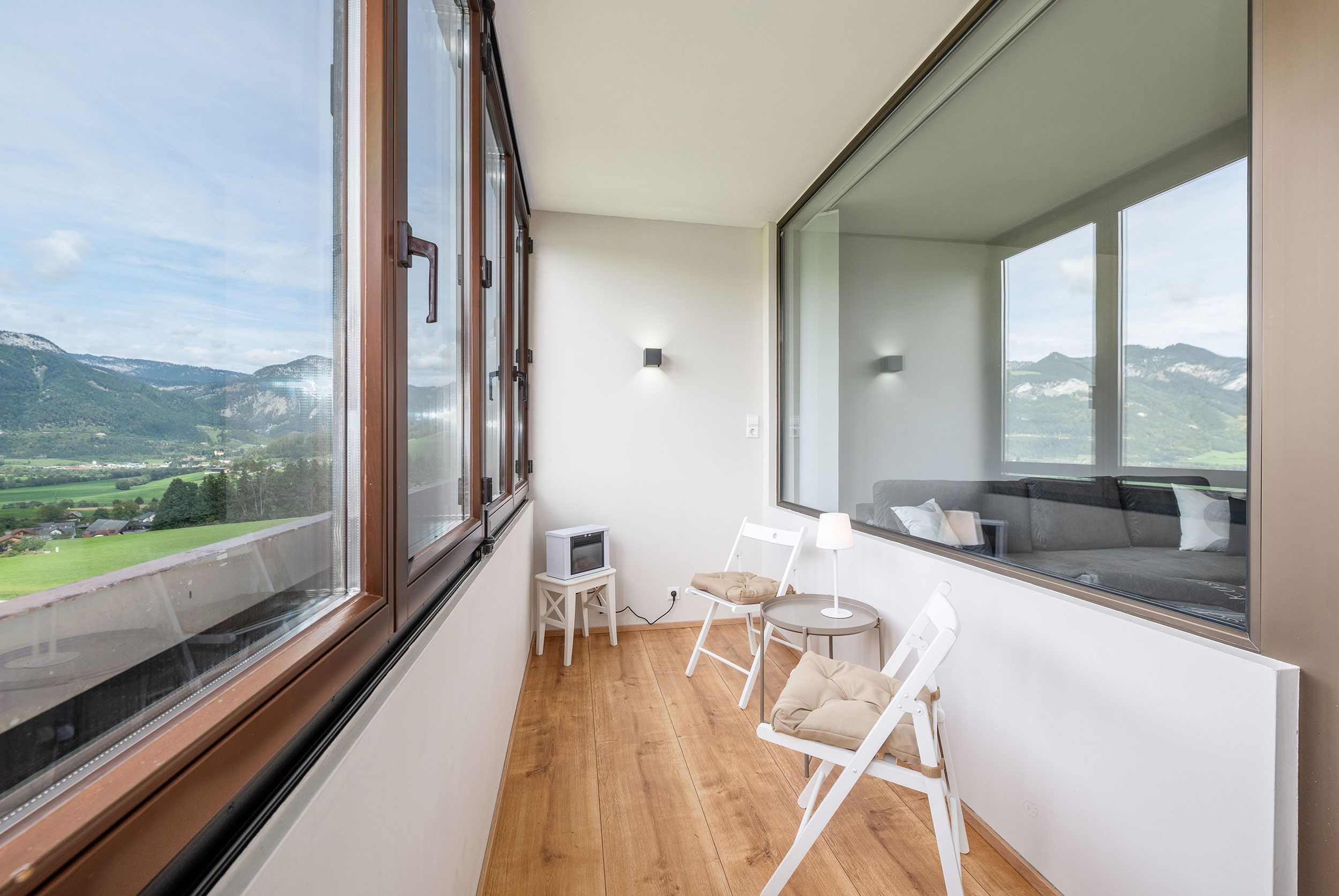 Un balcone con pavimento in legno, sedie bianche, un tavolino e un divano con cuscini.
