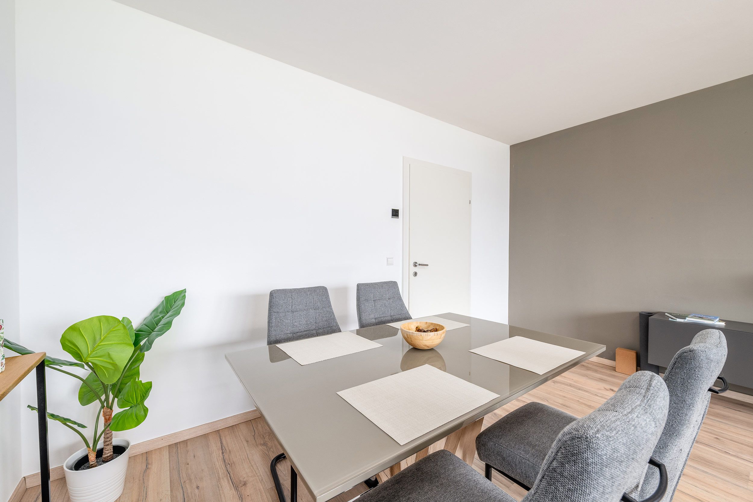 Una sala de reuniones moderna con una mesa de vidrio, sillas tapizadas en gris, una planta en maceta y una puerta al fondo.