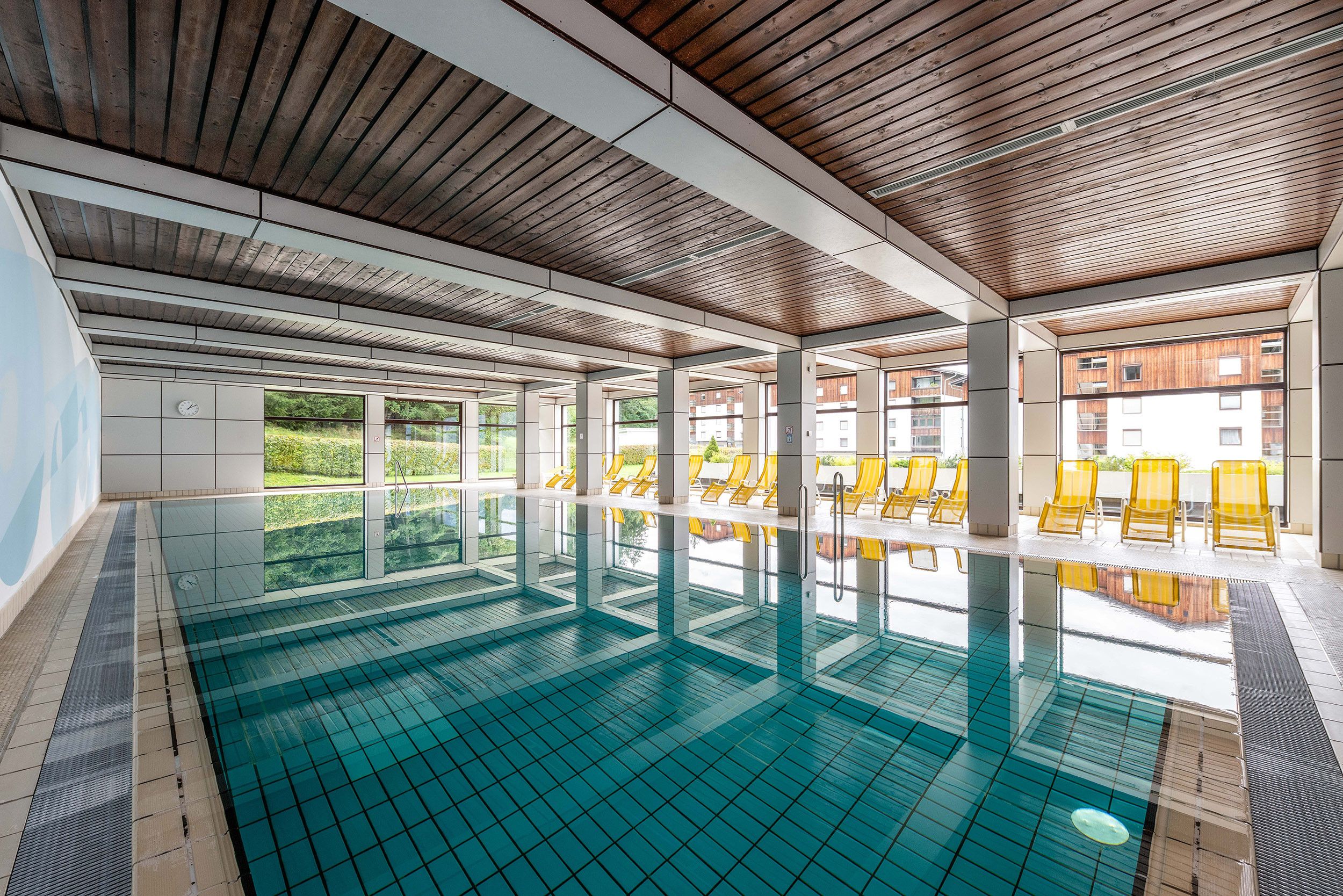 Una piscina interior con paredes de vidrio transparente, techo de madera y sillones de descanso amarillos.