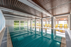 Una piscina interior con paredes de vidrio transparente, techo de madera y sillones de descanso amarillos.
