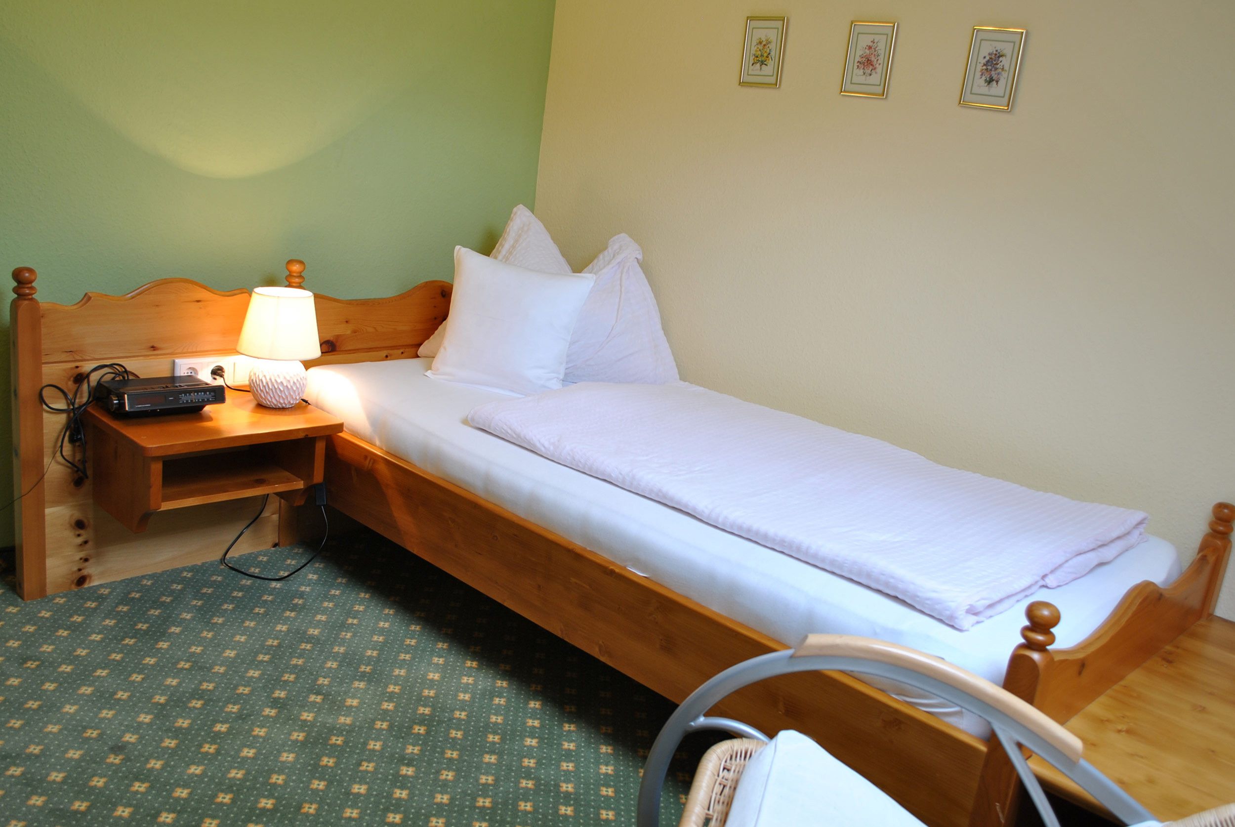 A single-bed room with a wooden headboard, a bedside table with a lamp, and a power strip. There are three framed pictures on the wall.