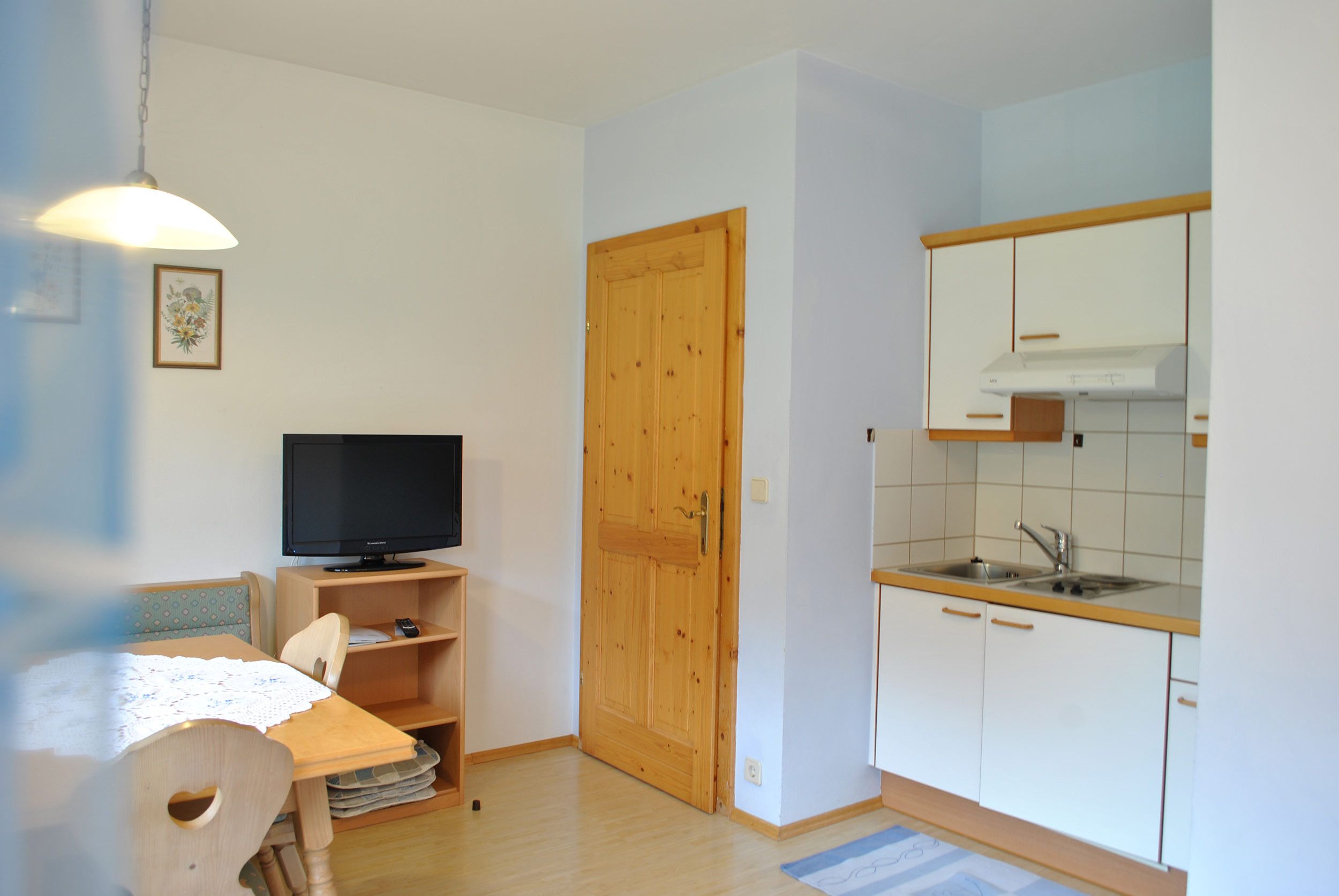 Una cucina compatta con una porta in legno, una televisione su un mobile basso e un piccolo angolo ristoro con tavolo e sedia.