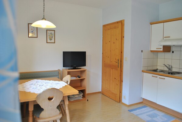 Une petite pièce bien rangée, avec une table à manger en bois, un téléviseur posé sur un meuble, une porte en bois et une kitchenette équipée d’un évier et de placards.