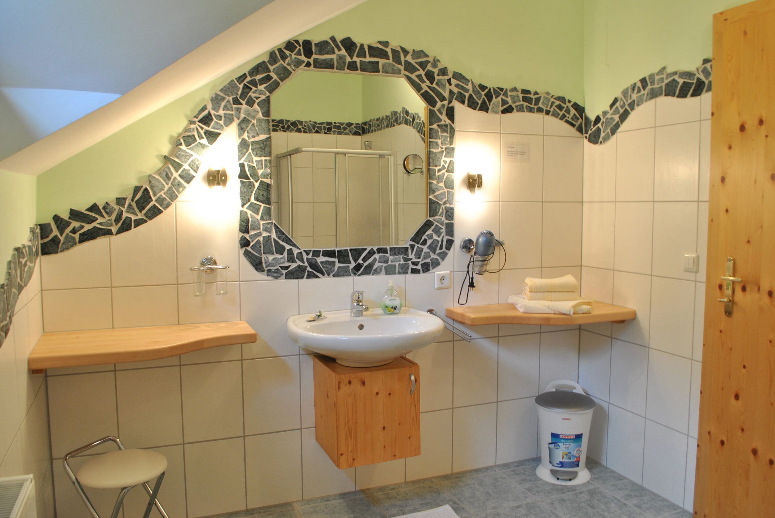 Un baño con un arco de mosaico de piedra sobre el espejo, un lavamanos con armario de madera, un perchero y un cubo de basura.
