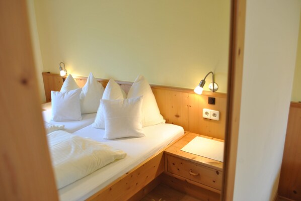 Una cama individual con ropa de cama blanca y almohadas, una mesita de noche con lámpara y un tocador de madera.