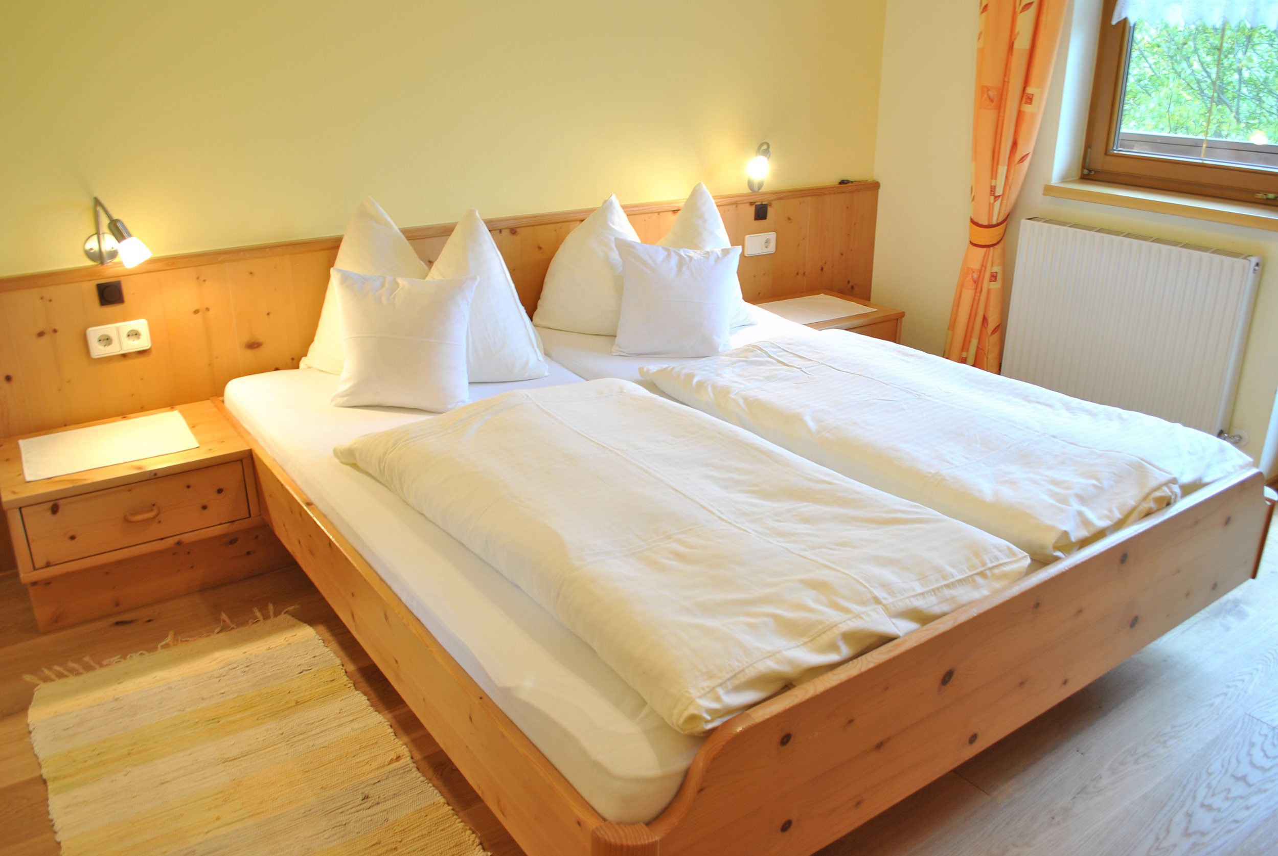 Cama doble con ropa de cama blanca, cabecera de madera, mesitas de noche y ventana con cortinas.