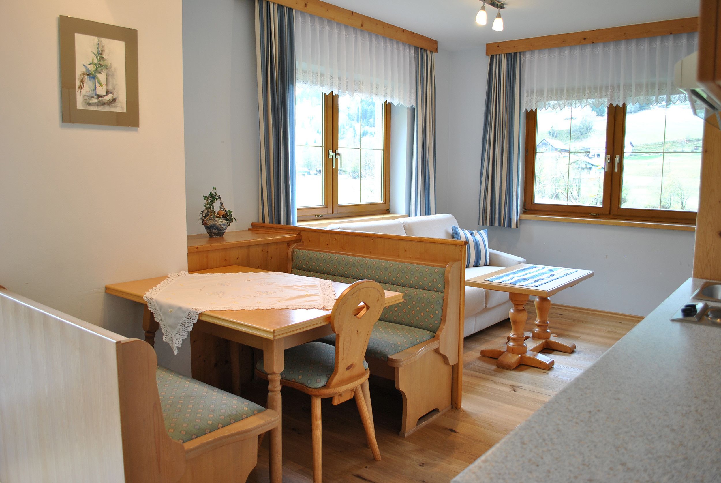 Una habitación acogedora con una mesa de madera, una banca y un sofá. Hay dos ventanas con cortinas y un cuadro en la pared.
